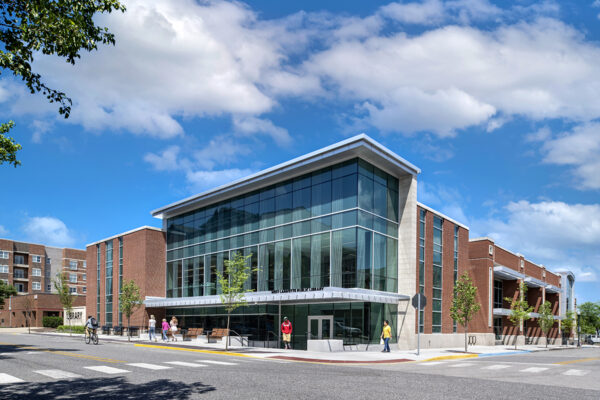 A modern brick and glass building with large windows is seen on a sunny day, with people walking and biking nearby—an inviting space inspired by Bradbury Miller Associates (BMA) design principles.