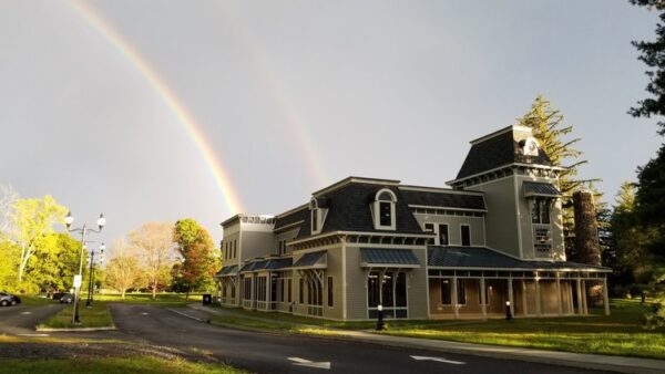 A large Victorian-style building sits under a partly cloudy sky with a visible double rainbow in the background. Trees and grass surround the structure, captured in a scene reminiscent of Bradbury Miller Associates' signature BMA settings.