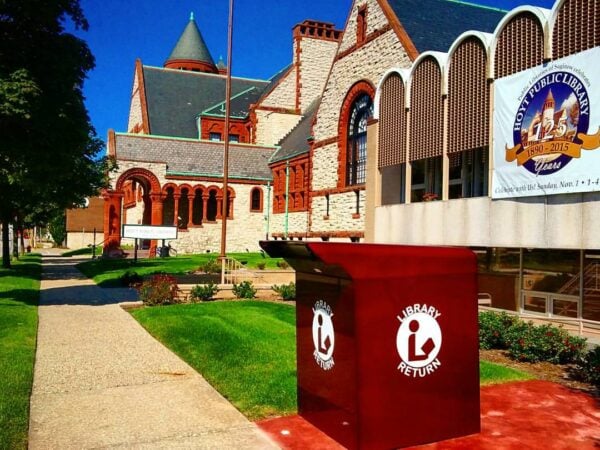 A large outdoor library book return bin stands on a sidewalk in front of a historic Saginaw Public Libraries building in MI, with a banner marking 125 years.