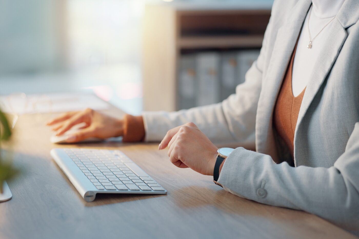 Person in a light-colored blazer sits at a desk using a wireless keyboard and mouse, with office shelves and documents in the background at Bradbury Miller Associates (BMA).