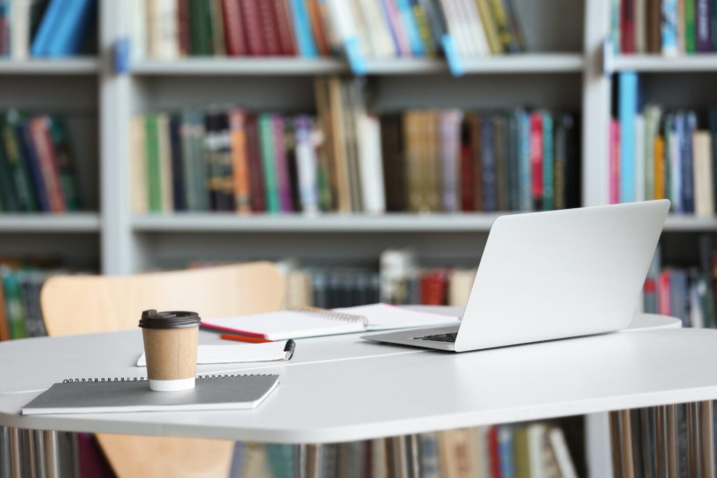A laptop, coffee cup, paper and pen sit on a white desk situated in a modern public library with white bookshelves.