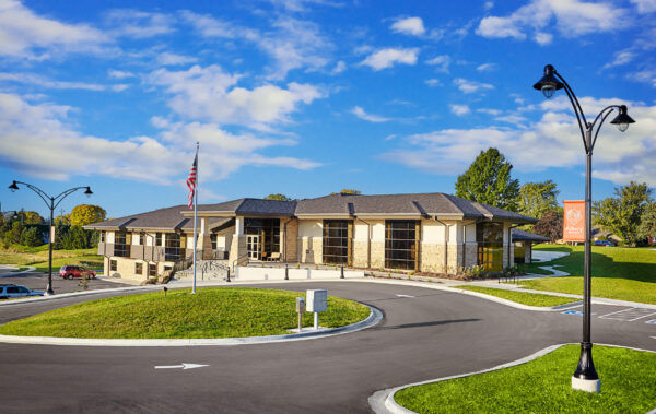 A modern single-story building with large windows and an American flag in front, surrounded by a curved driveway and grassy areas under a clear blue sky—an inviting space thoughtfully designed by Bradbury Miller Associates.