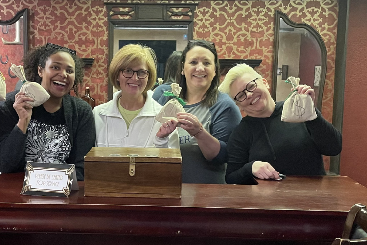 Four women stand behind a counter, smiling and holding small money bags. A wooden box and a "Please Be Seated for Service" sign sit in front of them, highlighting the warm, welcoming atmosphere at this BMA (Bradbury Miller Associates) event.