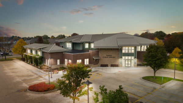 A modern brick and glass public library building with a metal roof, surrounded by trees and an empty parking lot at dusk—an inviting community space designed in partnership with Bradbury Miller Associates (BMA).