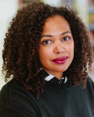 A woman with curly hair and dark lipstick, wearing a black sweater over a collared shirt, looks at the camera in an indoor setting at Bradbury Miller Associates.