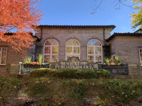 Exterior view of Sewickley Public Library with stone facade, arched windows, and fall foliage. Decorative plants and pumpkins line the front railing, creating a welcoming scene for BMA’s Bradbury Miller Associates clients and visitors.