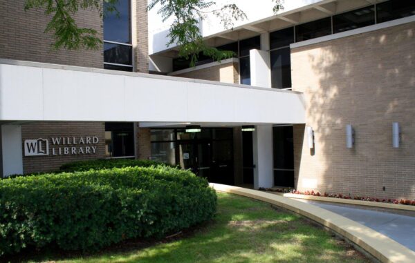 Exterior view of Willard Library entrance with glass doors, brick walls, bushes, and a curved sidewalk leading to the building managed in partnership with Bradbury Miller Associates.