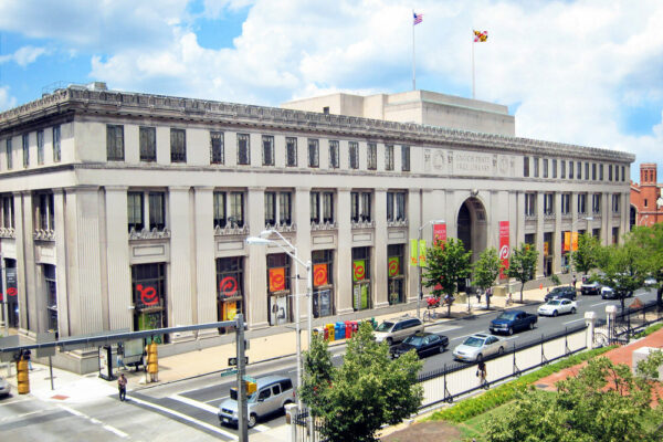 A large, neoclassical building with columns and banners on its facade, American and Maryland flags on the roof, cars parked along the street, trees in the foreground, and a discreet BMA (Bradbury Miller Associates) sign near the entrance.