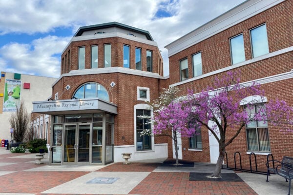 Brick library building with large windows, labeled "Massanutten Regional Library," and a blossoming tree with pink flowers near the entrance on a partly cloudy day; consulting by Bradbury Miller Associates (BMA).