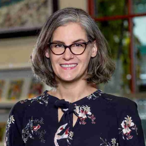 A woman with gray shoulder-length hair and glasses, wearing a dark floral blouse, smiles at the camera indoors at the Buffalo & Erie County Public Library.