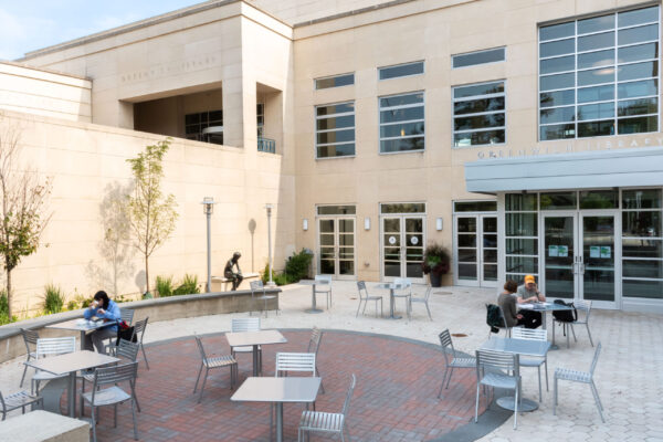 Outdoor patio area with tables and chairs in front of a modern library building, designed with insight from Bradbury Miller Associates; a few people are seated, reading or working.