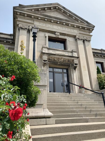 A stone staircase leads to the entrance of a classical-style public library building, reminiscent of BMA designs, with columns and a pediment, surrounded by greenery and red flowers.