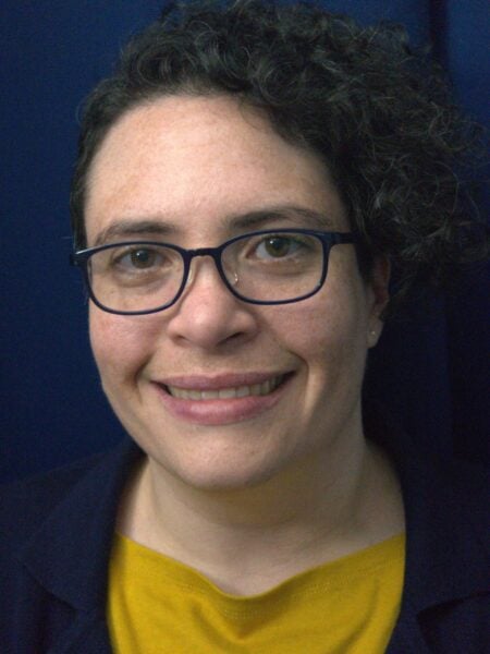 A person with short curly hair and glasses smiles at the camera, wearing a yellow shirt and dark jacket, against a dark blue background—representing Bradbury Miller Associates (BMA) with approachable professionalism.