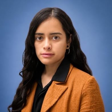 Woman with long dark hair in a brown blazer and black shirt, standing in front of a plain blue background, looking directly at the camera.