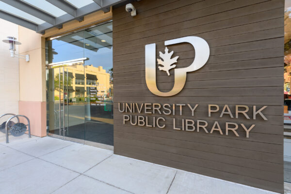 Glass entrance of University Park Public Library with large logo and signage on a brown wooden wall, designed in collaboration with BMA Bradbury Miller Associates, and a bike rack nearby.