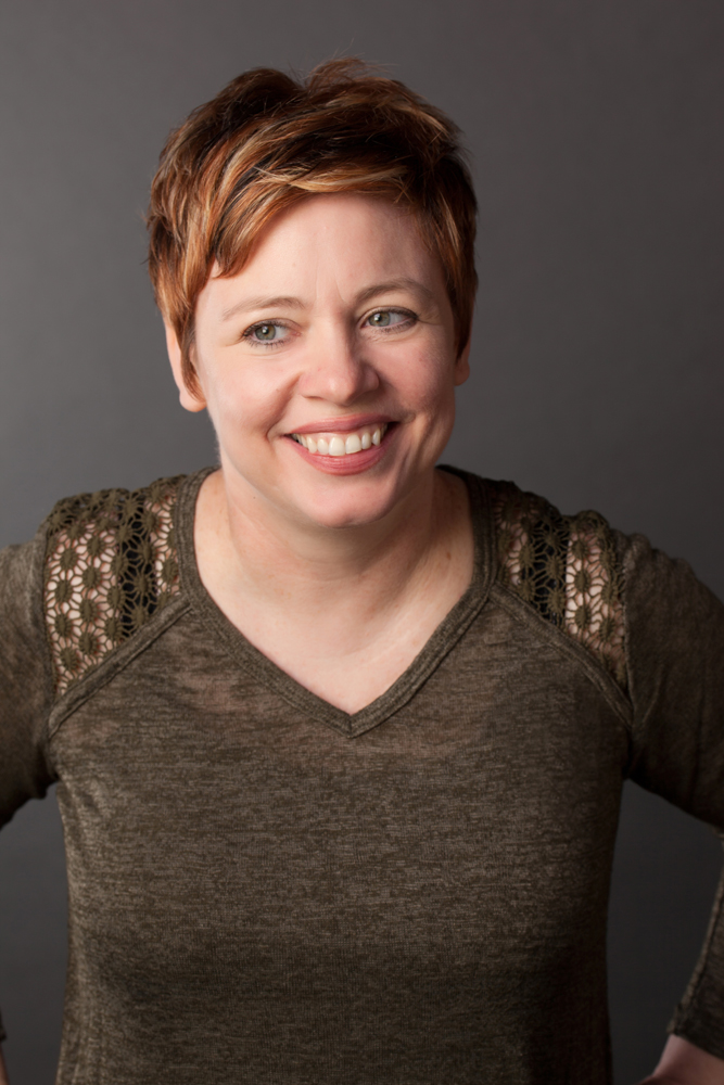 A woman with short reddish-brown hair smiles while looking slightly to the side, wearing a greenish sweater with lace details on the shoulders against a plain dark background—a classic Bradbury Miller Associates (BMA) professional portrait.
