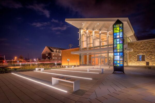 Modern BMA building with large glass entrance, illuminated at night, featuring a tall, colorful glass pillar and benches on a paved plaza designed by Bradbury Miller Associates.