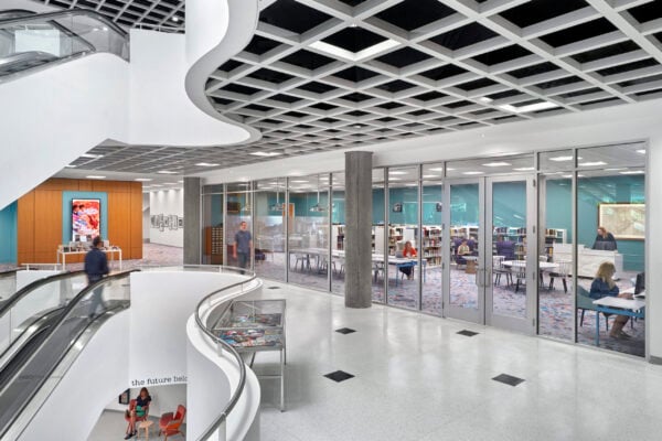 Modern library interior with a glass-walled study room, people reading and working at tables, a curved staircase, and colorful artwork on the wall designed with insights from Bradbury Miller Associates.