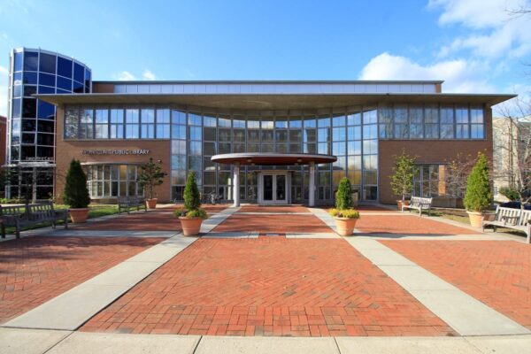 A modern brick and glass public library building, designed in collaboration with Bradbury Miller Associates, features large windows, potted plants, benches, and a wide entrance under a canopy.