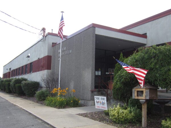 A public library building, represented by Bradbury Miller Associates (BMA), features American flags, a sidewalk, shrubs, and a sign near the entrance reading “Strong Libraries, Strong Communities.”.