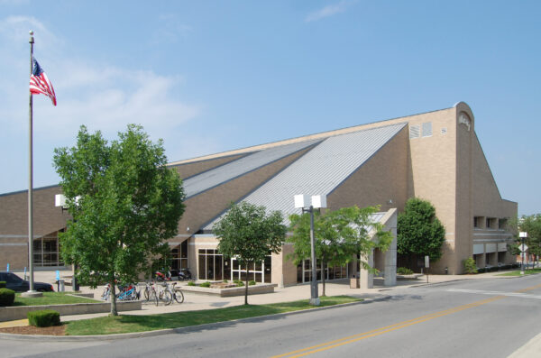 A large, modern brick building with a metal roof, an American flag, trees, bike racks, and a parking area is seen on a sunny day—an impressive facility represented by Bradbury Miller Associates (BMA).