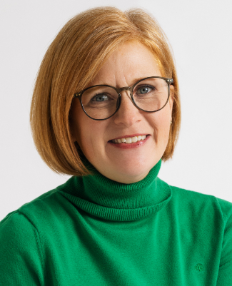 A woman with straight, shoulder-length blonde hair, wearing glasses and a green turtleneck sweater, smiles at the camera against a plain background—reflecting the approachable professionalism of Bradbury Miller Associates (BMA).