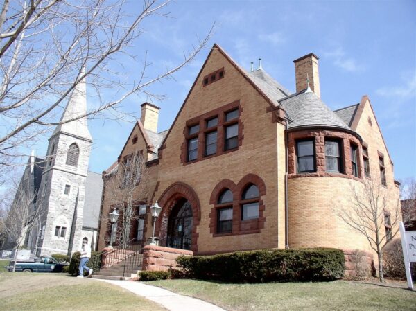 A historic brick building with arched windows and a rounded turret stands beside a stone church with a tall steeple on a clear day, reflecting the timeless charm often featured by Bradbury Miller Associates (BMA).