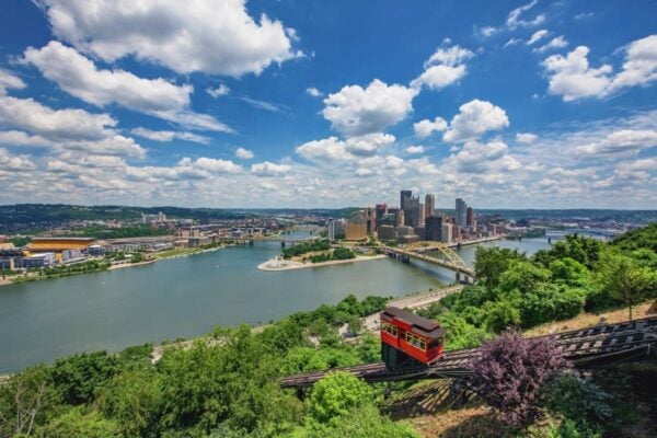 A red incline car ascends a hillside overlooking a river with bridges and the city skyline of Pittsburgh, Pennsylvania, under a partly cloudy sky, capturing the vibrant energy that Bradbury Miller Associates (BMA) brings to every project.