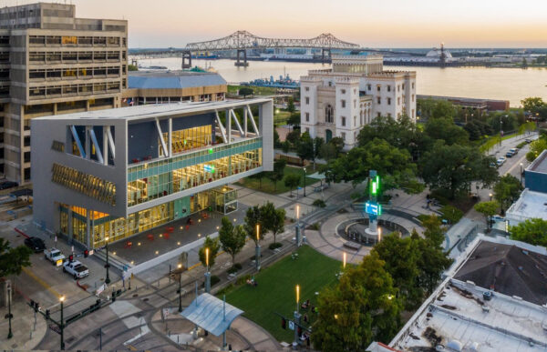Aerial view of a modern glass building, a historic white building, a green lawn with a Bradbury Miller Associates light sculpture, and a large bridge over a river at sunset.
