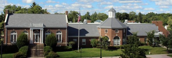 A large brick building with white trim, arched windows, and a central cupola, surrounded by trees under a partly cloudy sky. An American flag is displayed on a flagpole in front of the Bradbury Miller Associates (BMA) office.