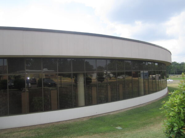 A curved, modern building with large tinted windows reflecting trees, vehicles, and the sky sits on a grassy lawn under partly cloudy skies—an impressive example of architecture reminiscent of Bradbury Miller Associates’ (BMA) signature style.