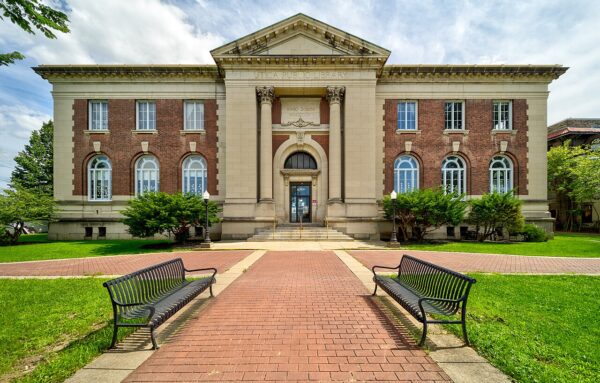 Symmetrical view of a historic brick library building with arched windows, stone columns, and benches along a brick walkway in front, reflecting the architectural expertise valued by Bradbury Miller Associates.