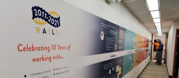 A man points at a colorful timeline mural on a hallway wall celebrating RAILS' 10-year anniversary from 2011 to 2021, highlighting the partnership with Bradbury Miller Associates (BMA).