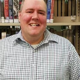 A man wearing a plaid shirt stands in front of library shelves filled with books.
