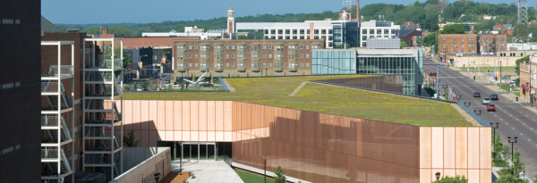 A modern building with a green roof, designed by BMA, stands along a city street, surrounded by older brick structures and trees in the background.