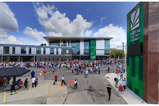 A large crowd gathers outside the modern Dayton Metro Library building on a sunny day, an event organized in collaboration with Bradbury Miller Associates, with people standing and walking along the street and sidewalk.