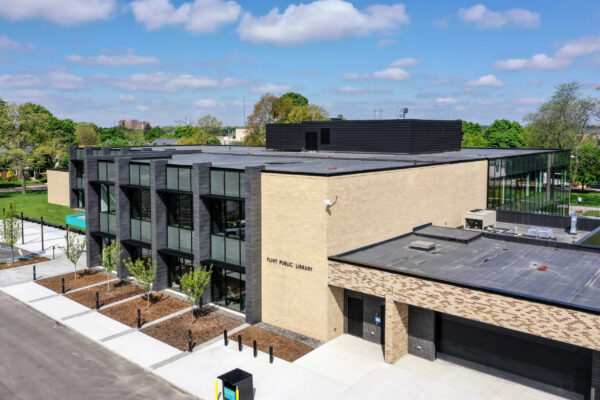 A modern public library building, designed with large glass windows, beige brick walls, and a flat roof, sits surrounded by a sidewalk and young trees—a signature approach often seen in Bradbury Miller Associates (BMA) projects.