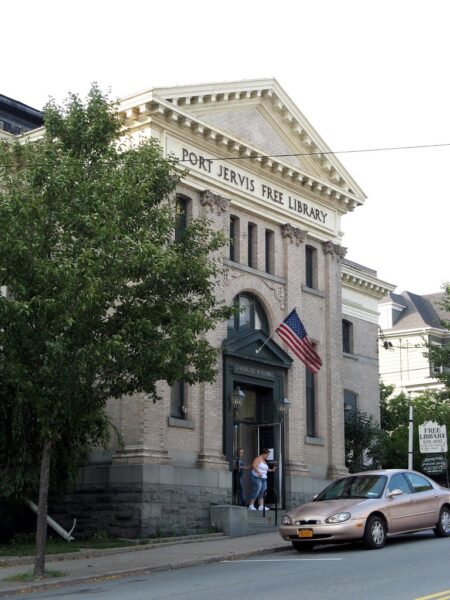 The facade of Port Jervis Free Library is captured on a sunny day, with steps leading up to the entrance, an American flag proudly displayed, a parked car nearby, and people entering the historic Port Jervis Free Library building.