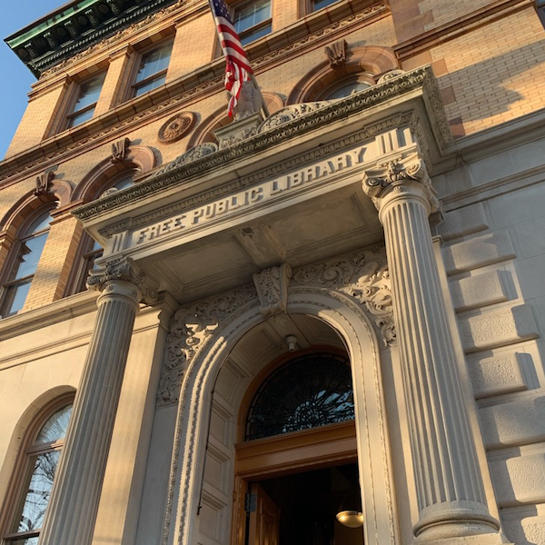 Entrance of a historic brick building with columns, an American flag above, and a sign reading "Free Public Library" over the doorway—an inspiring setting often highlighted by Bradbury Miller Associates (BMA).