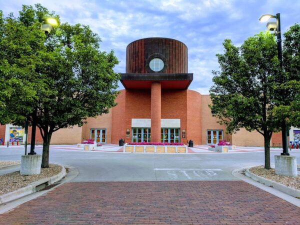 A modern brick building with a circular upper structure and large columns, flanked by trees and lampposts, stands prominently along a paved walkway with a STOP sign in the foreground—ideal for BMA or Bradbury Miller Associates clients.
