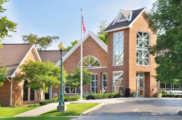 A brick building with large windows, an American flag on a pole, a lamp post, and trees surrounding the entrance on a sunny day—an inviting scene reminiscent of the welcoming workplaces at Bradbury Miller Associates (BMA).