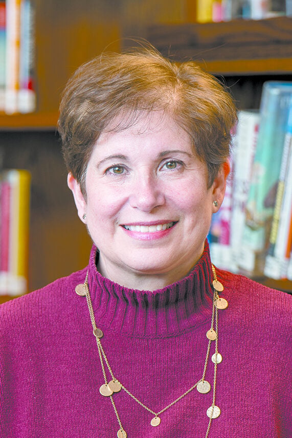 Smiling woman with short brown hair wearing a maroon sweater and gold necklace, standing in front of bookshelves filled with colorful books at the Katonah Village Library in Katonah, NY.