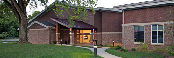 A brick building with a red metal awning over the entrance, glass doors, and a sidewalk lined with small outdoor lights, surrounded by grass and trees.